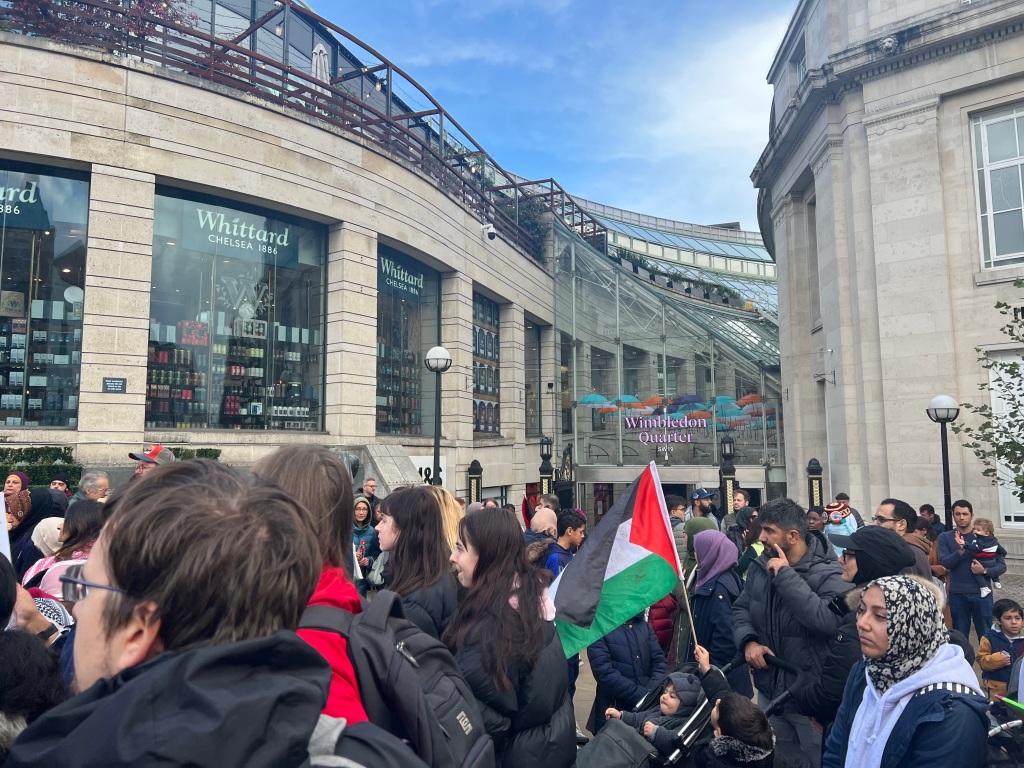 Image showing vigil crowd outside Wimbledon Quarter shopping centre with a Palestine flag raised. People are looking towards the centre of the crowd.